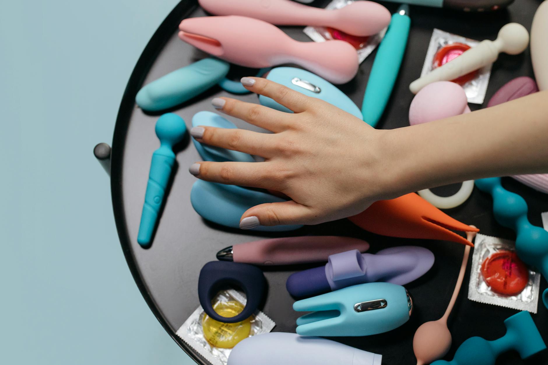 Mão alcançando uma variedade de brinquedos sexuais coloridos sobre uma mesa