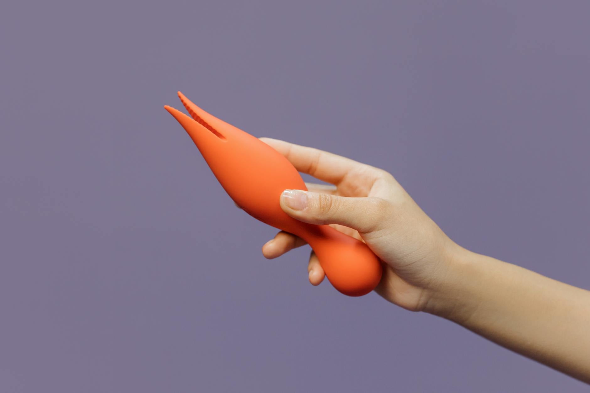 Mão segurando um vibrador laranja contra fundo minimalista roxo, mostrando sensualidade moderna.
