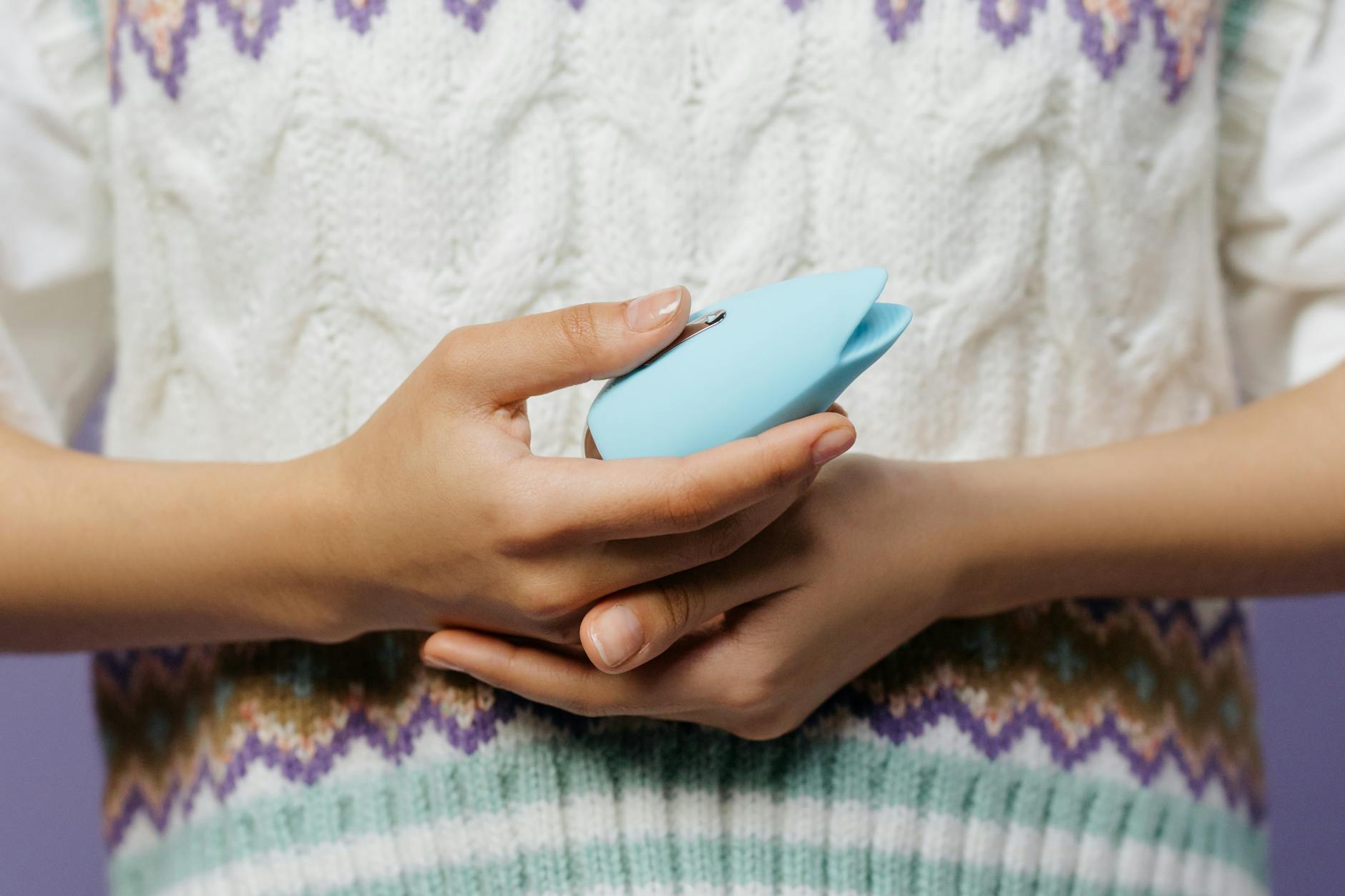 Foto aproximada de mãos segurando um vibrador azul contra um suéter de tricô.